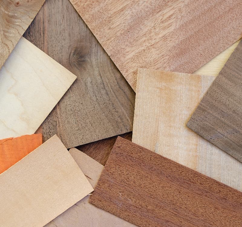 A flat lay arrangement of various wood veneer samples in different tones and grain patterns, ranging from pale birch and light oak to warm mahogany and dark walnut, overlapping each other on a wooden surface.